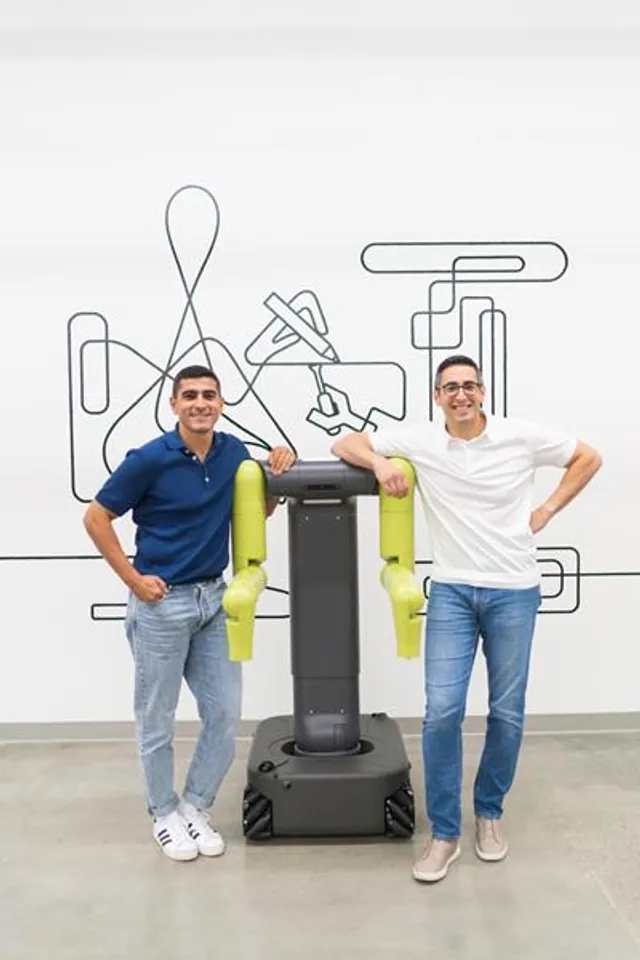 Maven Robotics co-founders Hamza and Khalid Derbas stand on either side of the company's wheeled, dual-arm industrial robot in an office space, the robot's dark mobile base topped by two articulated arms ending in lime-green gripper housings, a line-art wall mural behind them.