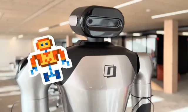 A humanoid robot platform used to demonstrate Robostral Navigate, showing its dark rounded head unit with dual front-facing cameras and silver-and-black torso, photographed indoors in an office setting with a small Mistral mascot sticker on its shoulder