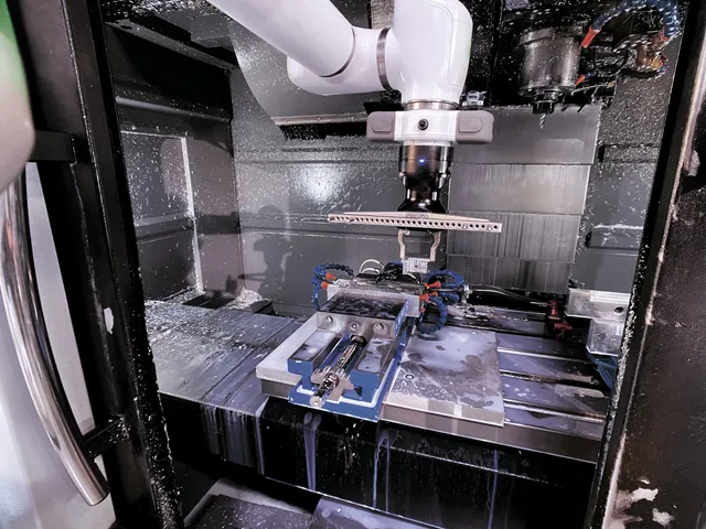 A white OB7 collaborative robot arm reaching into an active CNC milling machine coated in coolant spray, tending a vise-held workpiece