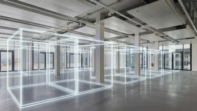 An empty industrial interior space with concrete floors and support columns, marked out with illuminated white wireframe cube outlines suspended from the ceiling, inside the NEURA Gym RWTH Aachen facility.