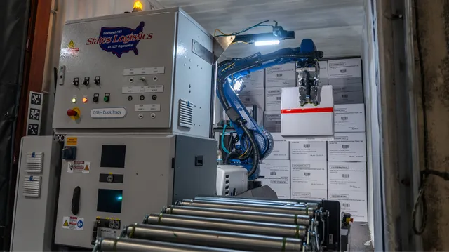 A KUKA robotic arm unloading boxed cases from inside a semi-trailer next to a States Logistics control panel, with a roller conveyor in the foreground.