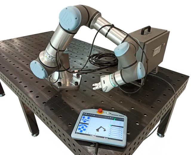 A Universal Robots UR16e collaborative robot arm mounted on a fixturing table, with its gray controller box behind it and a teach pendant showing the robot's joint positions and an emergency-stop button in the foreground.