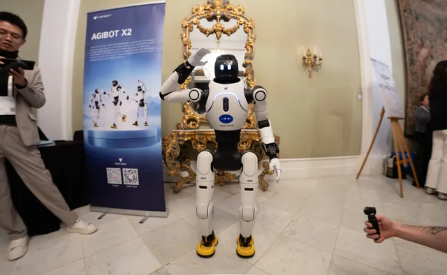 AGIBOT's X2 humanoid robot, a white and dark bipedal robot with a rounded head and slim torso, standing on display at a trade-show booth.