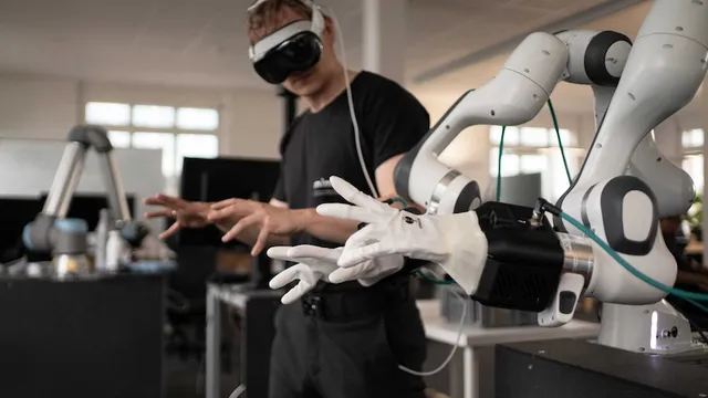 A man wearing a VR headset and white glove-based teleoperation controllers, reaching toward a white-and-black six-axis robotic arm fitted with a dexterous gripper, in an office/lab setting.