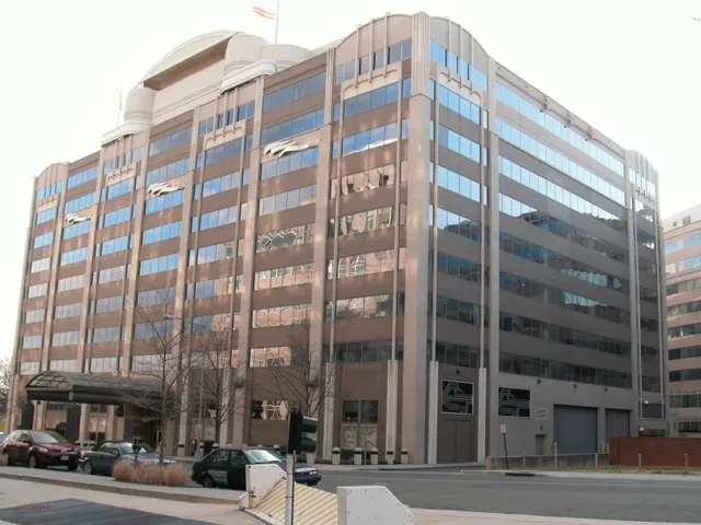 Exterior street-level view of the FCC headquarters building in Washington, D.C. — an art-deco-influenced modern office building with cars parked out front and bare winter trees.