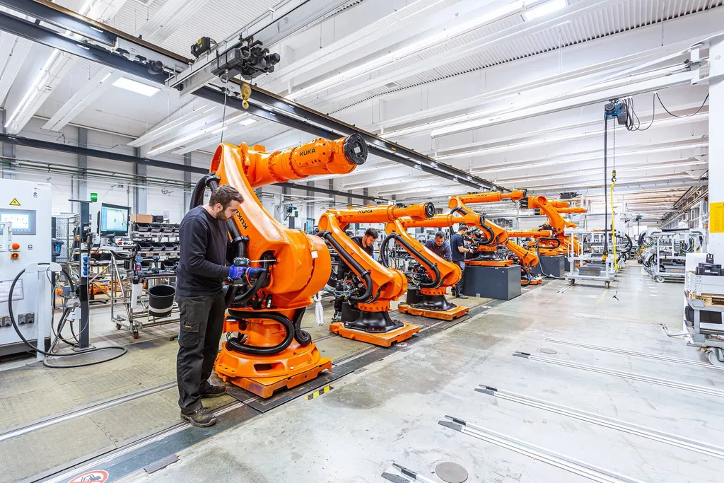 A technician fits cabling to one of a row of orange KUKA six-axis robot arms on a factory assembly line, with overhead crane rails and workstation monitors visible behind.