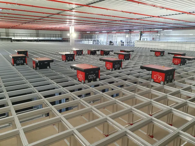Numbered red-and-black AutoStore robots sit atop an aluminum storage grid in a warehouse, with rows of storage bins visible beneath the grid cells.