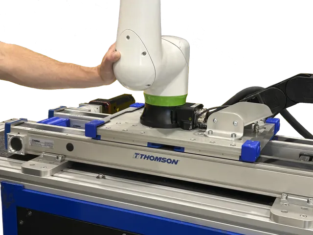 A technician's hand positions a white and green FANUC CRX collaborative robot arm mounted on a blue-and-silver Thomson Movotrak linear rail unit, with THOMSON branding visible on the rail.
