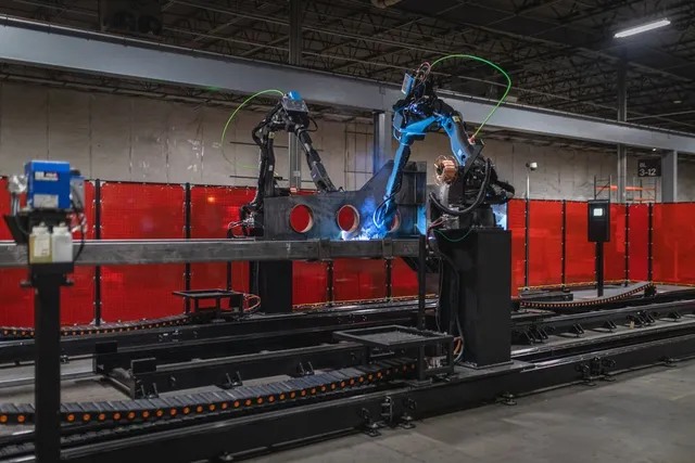 Two robotic welding arms on a rail-mounted gantry system welding a large steel ship structure inside a fabrication bay, with red safety curtains and visible weld sparks.