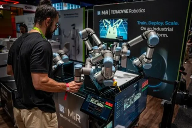 A man in a black t-shirt physically guides a Universal Robots cobot arm at a trade-show booth, capturing real-world motion data, with a second cobot arm and a monitor beside him under a Teradyne Robotics banner reading 'Train. Deploy. Scale. On an industrial platform.'