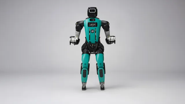 A teal-and-black bipedal humanoid robot with human-like legs stands facing forward against a plain gray studio backdrop, its chest display reading '1024 AUTO' and three-fingered grippers hanging at its sides.
